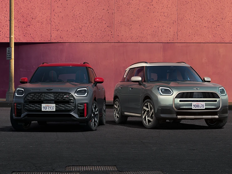 Kuva edestä MINI Countryman JCW:stä Legend Grey -värissä ja punaisella katolla sekä vieressä olevasta MINI Countrymanista Smokey Green -värissä ja Vibrant Silver -katolla.