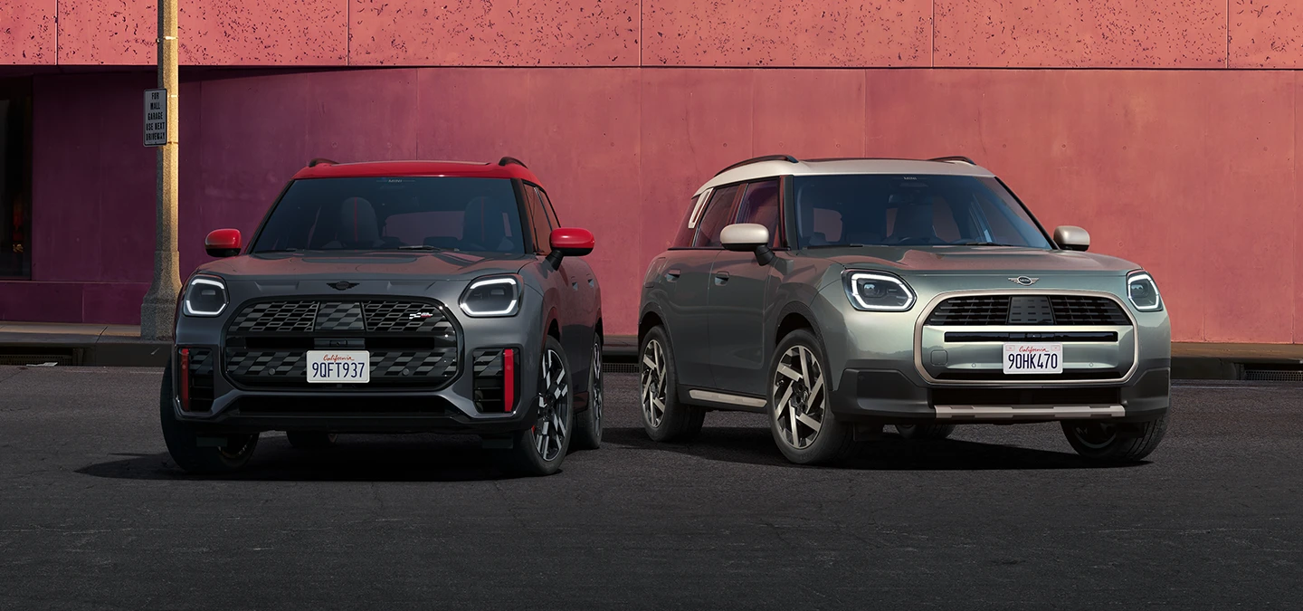 Kuva edestä MINI Countryman JCW:stä Legend Grey -värissä ja punaisella katolla sekä vieressä olevasta MINI Countrymanista Smokey Green -värissä ja Vibrant Silver -katolla.