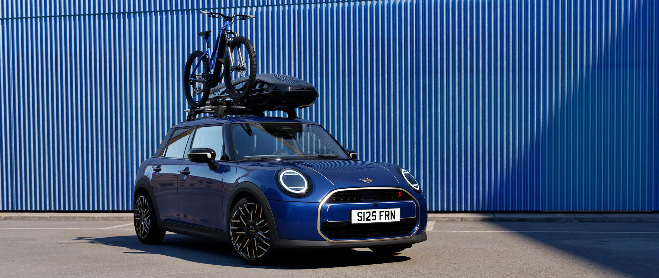 Kuva edestä Indigo Sunset Blue -värisestä MINI Acemanista MINI-kattopyörätelineellä, jossa on maastopyörä ja MINI-kattoboksi 320.