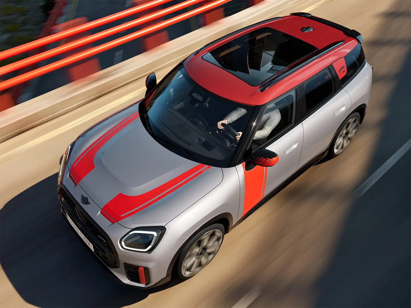 MINI Countryman John Cooper Works, väri Melting Silver, Chili Red ‑väriset peilinkuoret, sivu- ja konepeltitarrat, katto ja siivekkeet, ajaa kaupungilla katua pitkin – näkymä edestä.
