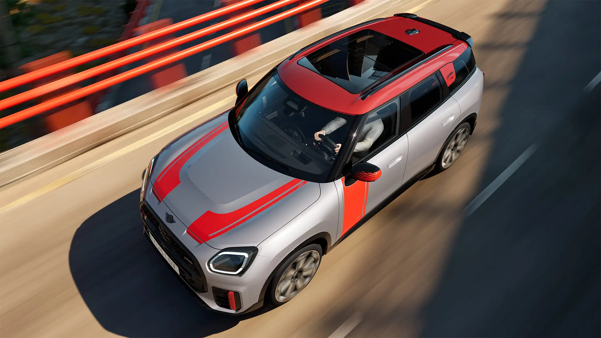 MINI Countryman John Cooper Works, väri Melting Silver, Chili Red ‑väriset peilinkuoret, sivu- ja konepeltitarrat, katto ja siivekkeet, ajaa kaupungilla katua pitkin – näkymä edestä.