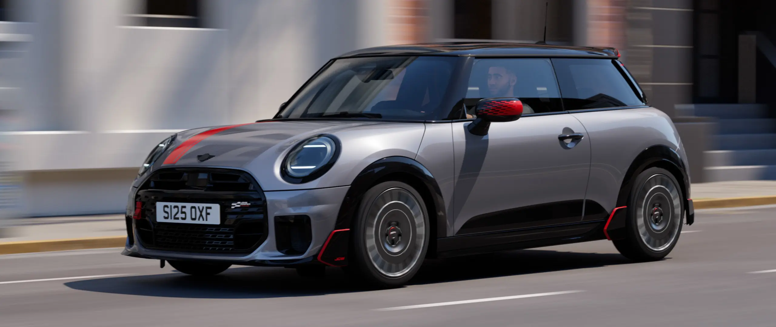MINI Cooper, 3-ovinen, väri Melting Silver, Chili Red ‑väriset John Cooper Works ‑peilinkuoret ja konepeltitarra, musta kattospoileri ja siiveke, ajaa kaupungilla katua pitkin – näkymä edestä.