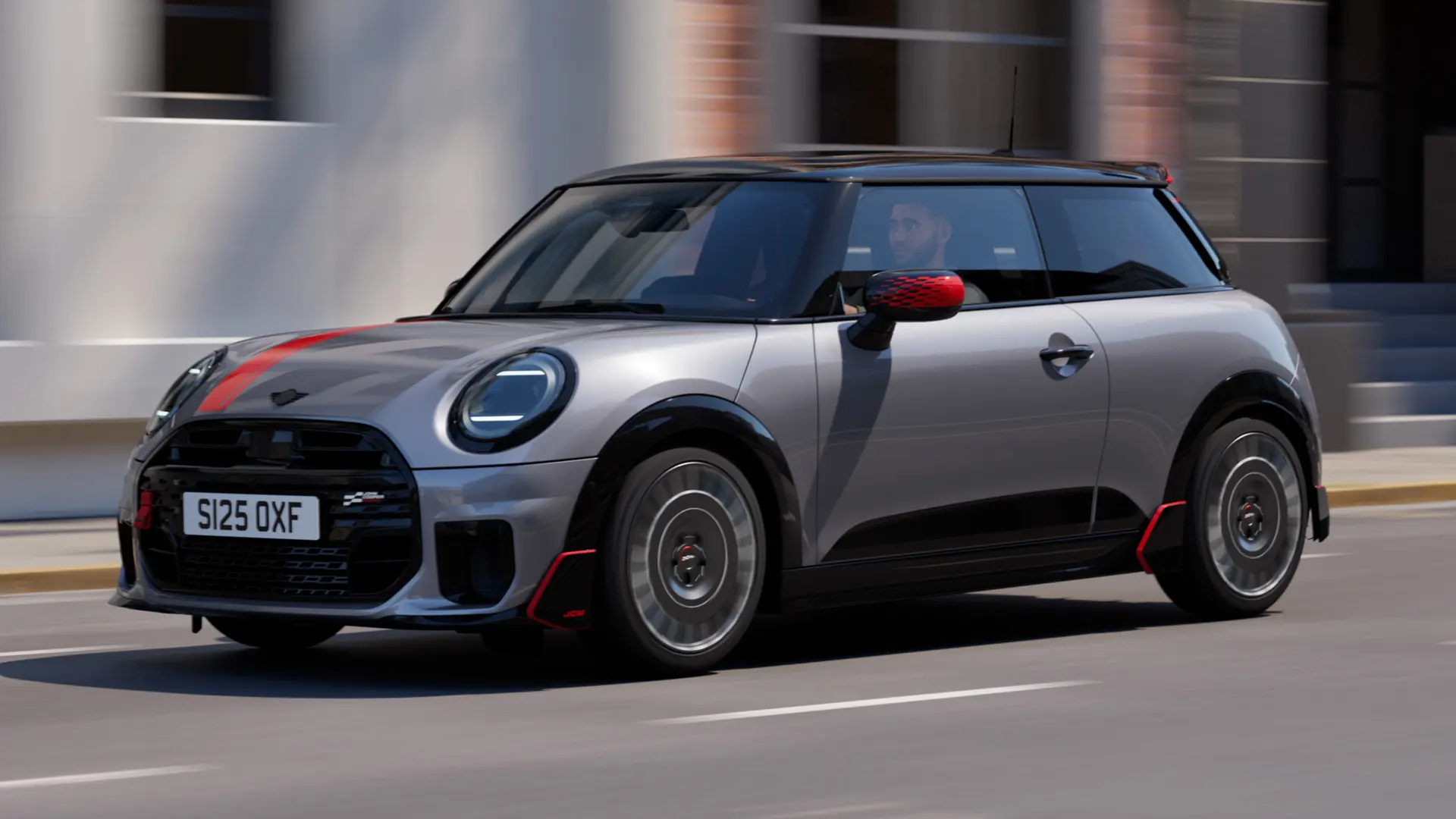 MINI Cooper, 3-ovinen, väri Melting Silver, Chili Red ‑väriset John Cooper Works ‑peilinkuoret ja konepeltitarra, musta kattospoileri ja siiveke, ajaa kaupungilla katua pitkin – näkymä edestä.