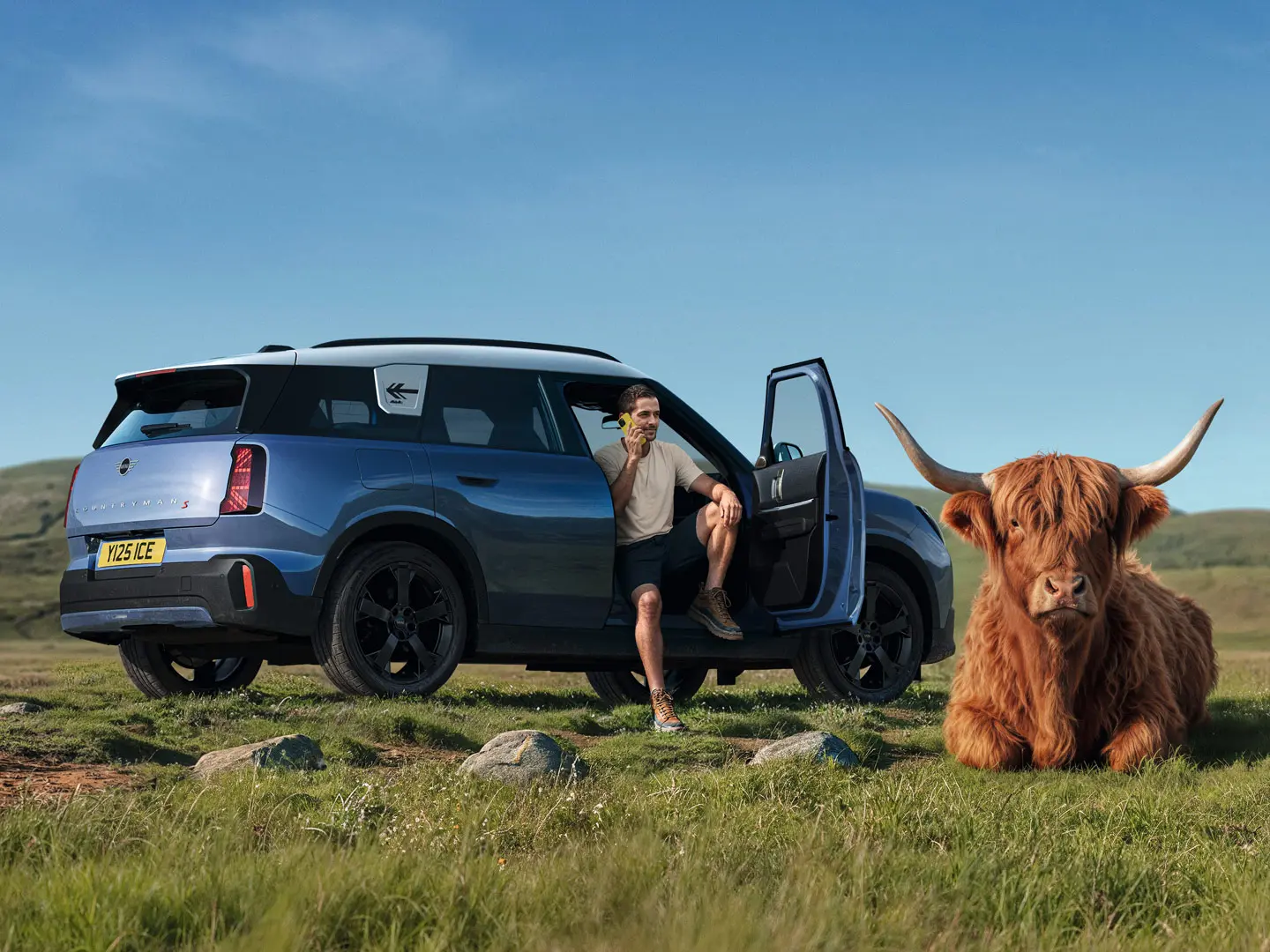 Mies puhuu MINI Countryman S -autonsa ulkopuolella MINI Liikkuvuuspalveluun MINI Appin kautta, ja lähistöllä loikoilee ylämaan härkä.